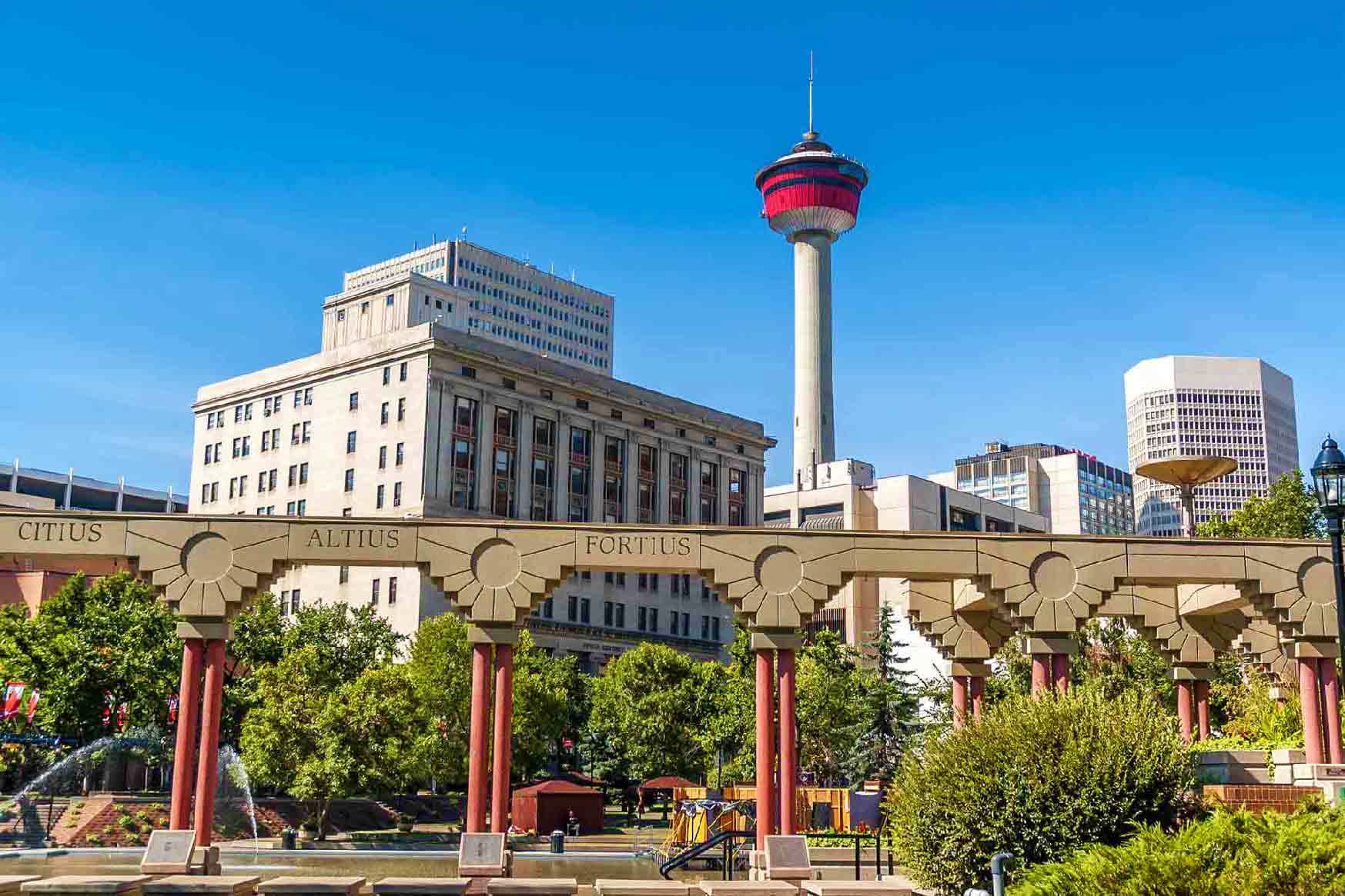 Calgary skyline with Bow Tower and historic downtown buildings framed by Eau Claire bridge with red columns and Latin inscrip